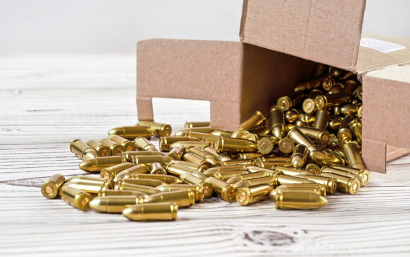 Yellow brass gun ammo spilled from paper carton box on white boar desk - close up.