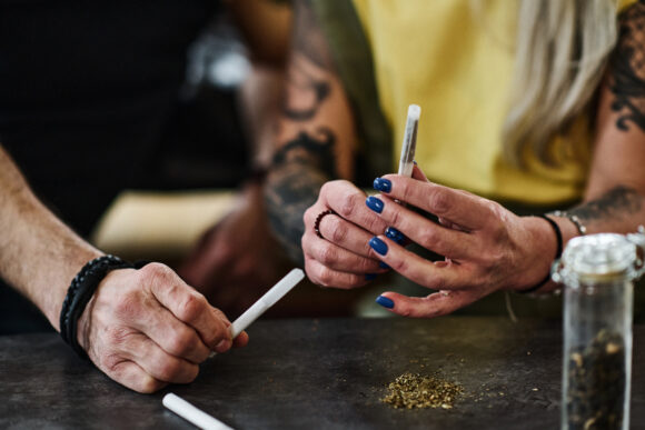 Unrecognizable mature tattooed woman and her boyfriend sitting at table rolling joints with marijuana
