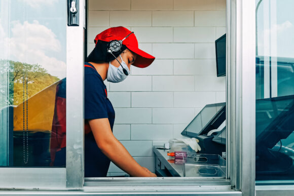 Bangkok, Thailand - May 28, 2020 : fast food cashier in drive thru service waring hygiene face mask to protect coronavirus pandemic or covid-19 virus outbreak working on counter at the station.