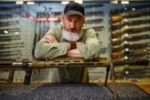 Bearded male hunter at the counter with powerful rifle in gun store. Weapon shop interior, ammo and ammunition assortment, firearms choice, shooting hobby and lifestyle