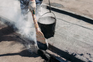 Worker repairs the roof with molten tar from a bucket with a broom. Roof repair tar.