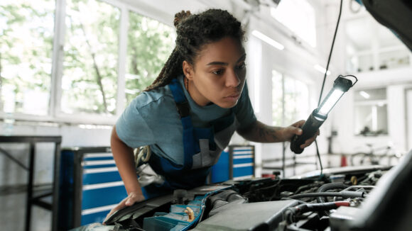 Young african american woman, professional female mechanic examining under hood of car with torch at auto repair shop. Car service, maintenance and people concept. Selective focus. Web Banner