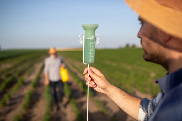 Male farmer standing in field holding rain gauge in forefront. Male farmer working in field spraying plants with portable hand sprayer.