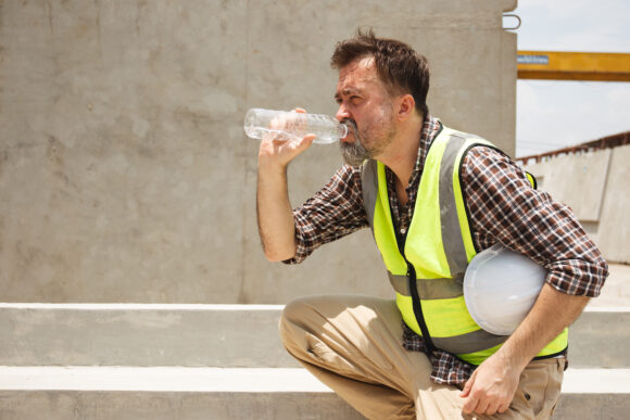 Dangerous from heat wave during working outside the building. Caucasian male construction worker resting and drinking water from bottle in a very hot day. Heat stroke health problem.