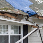 Residential house crushed by fallen trees and tree limbs during severe winter storm with strong winds. Tarp is placed on the damaged rooftop area as a temporary measure before proper roof repairs.